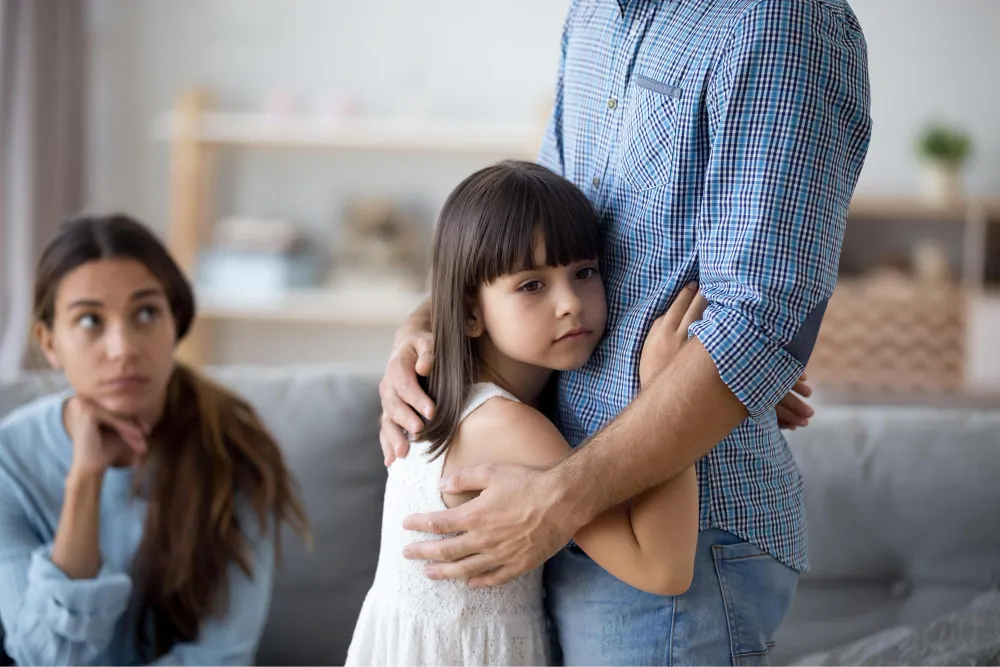 Menina abraça o pai, enquanto a mãe observa com preocupação sobre a guarda.